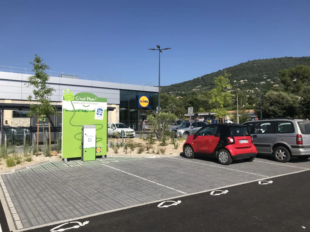 Borne de recharge pour véhicule électrique installée sur un parking avec dalles perméables O2D, en extérieur avec des voitures stationnées autour.